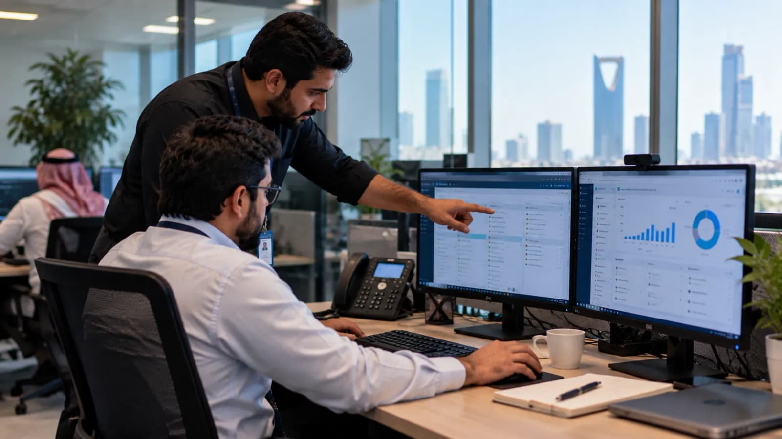 Two colleagues at a two-monitor workstation reviewing a customer record list and a sales dashboard